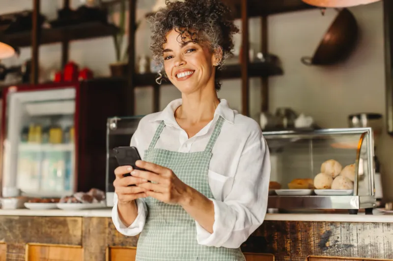 small business owner using a smartphone in her cafe mature businesswoman smiling at the camera while standing in front of the counter successful entrepreneur communicating with her customers online