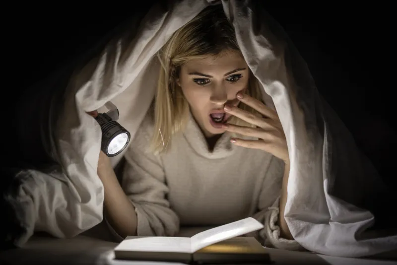 beautiful young woman reading book under bed cover and holding a flashlight