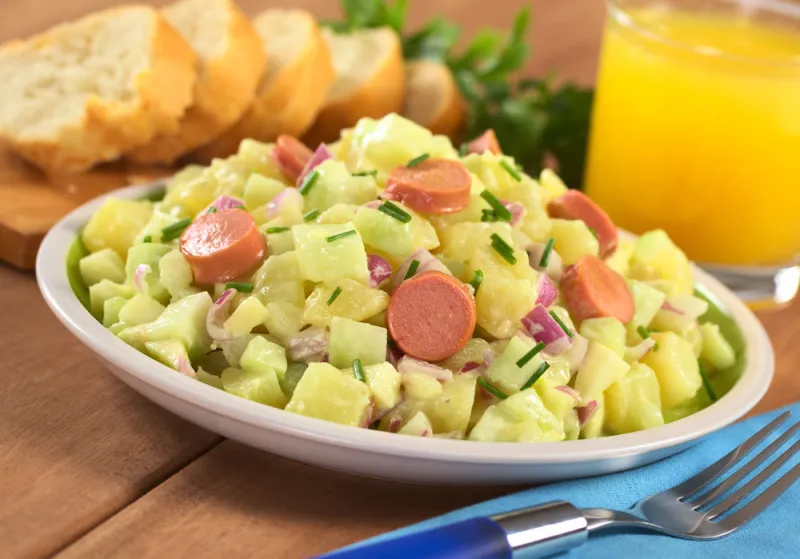 fresh potato salad made of potato, cucumber, red onion and chives with a mayonnaise dressing and sausage slices with orange juice and baguette slices in the back (selective focus, focus one third into the salad, on the sausage slice in the middle)