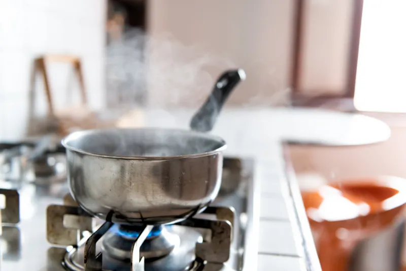 closeup of vintage tiled gas stove top with tiles white countertop and stainless steel pot and steam cooking with blue flame in retro kitchen