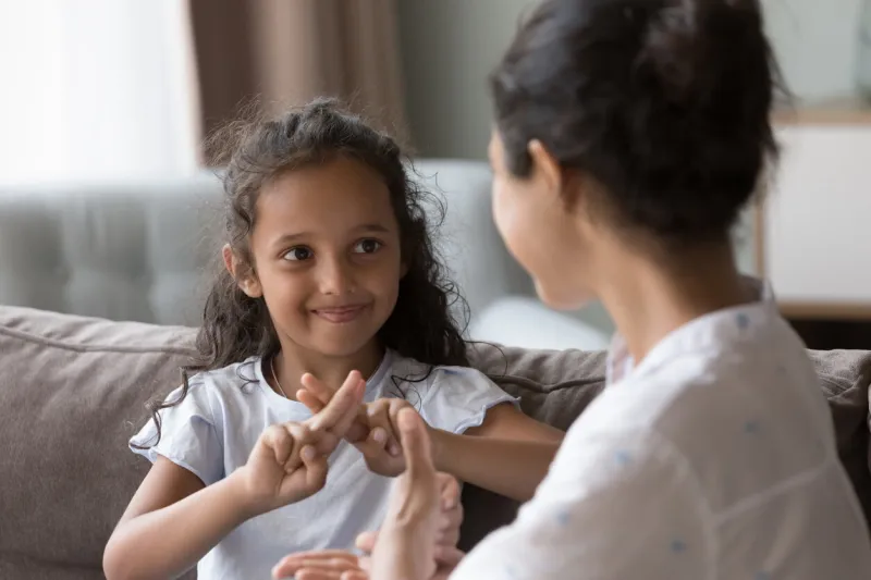 happy cute indian kid girl talking to mother, using hands, fingers, speaking sign languages, sitting on sofa at home female teacher, therapist teaching child with deafness to communicate