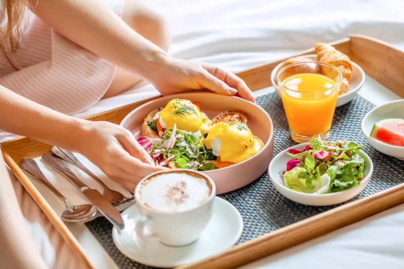 breakfast in bed served with cup of coffee, salad, fresh fruits and eggs benedict on wooden tray woman hands holding plate with fresh food room service in hotel