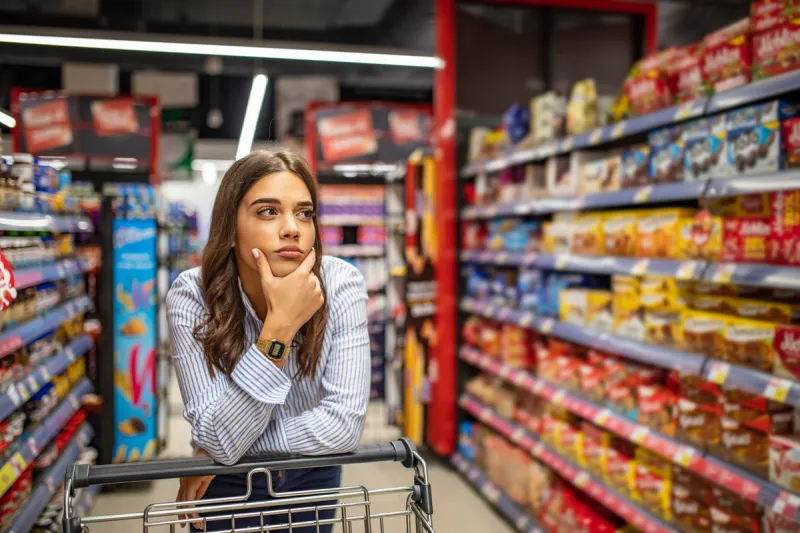 young woman staying confused in grocery store confused woman doesnt know what to buy young woman with a shopping cart at supermarket looking at supermarket shelf