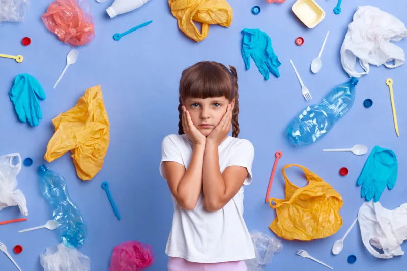 indoor shot of depressed little girl wearing white t shirt posing against blue wall and much plastic garbage around, keeps palms on cheeks, asks stop destroying our planet