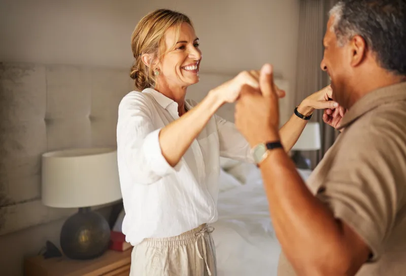 older interracial couple dancing in a hotel bedroom together on a romantic holiday senior hispanic man and caucasian woman smiling and playing at home, relaxing and content