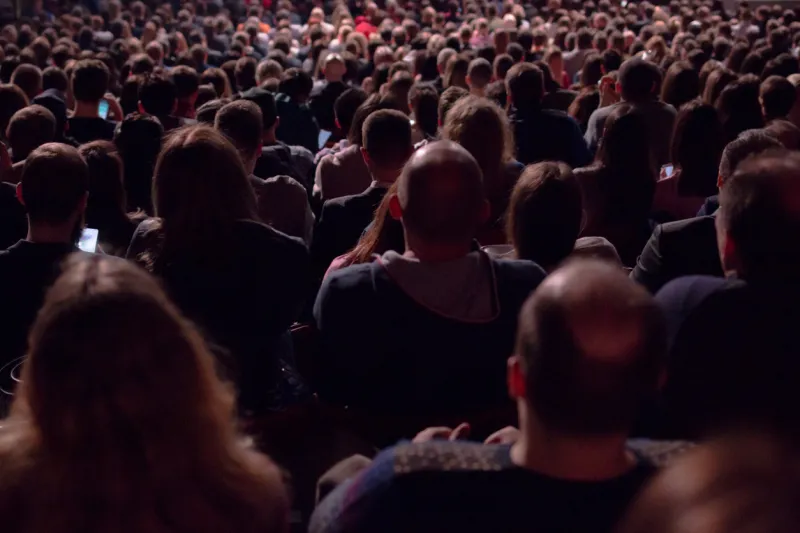 in the dark hall there is a view from the back of a crowd of hundreds of people sitting and watching the screen in a movie theater or performing on stage concept for articles