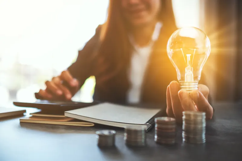 businesswoman holding a lightbulb with coins stack on table, saving energy and money concept
