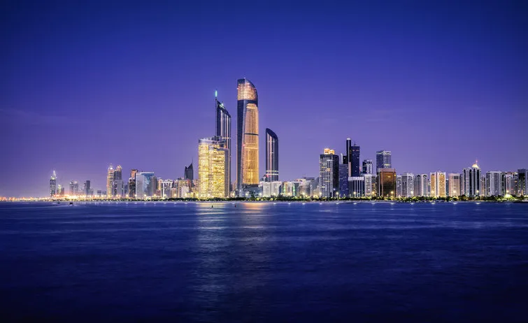 the skyline of abu dhabi at sunset, taken from the distance over water all of the buildings have their lights on, and the large towers in the middle are the central market towers the sea is very tranquil, and the fading light lingers behind the city