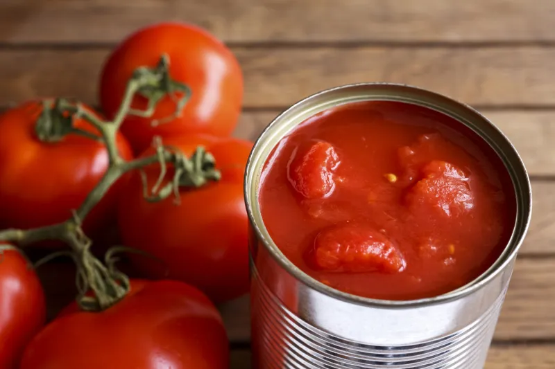 open tin of chopped tomatoes with whole fresh unfocused tomatoes behind wood surface