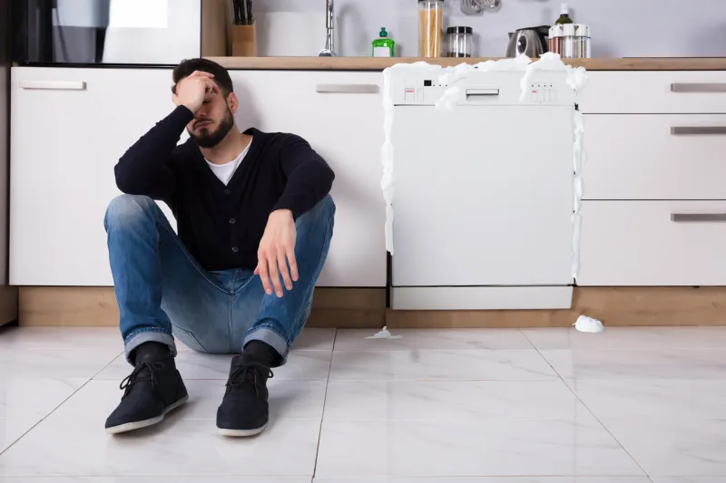 upset man sitting next to dishwasher with foam coming out in kitchen