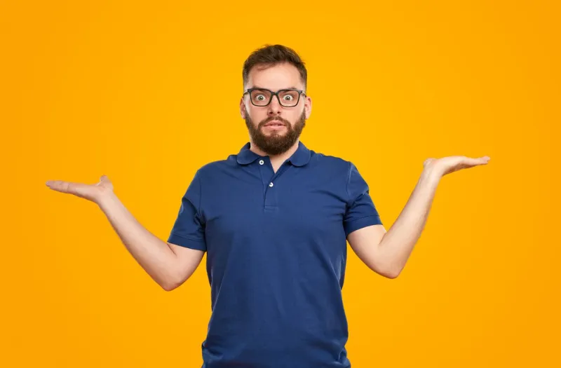 amazed bearded male in casual outfit and eyeglasses keeping palms up while making choice and looking at camera against yellow background