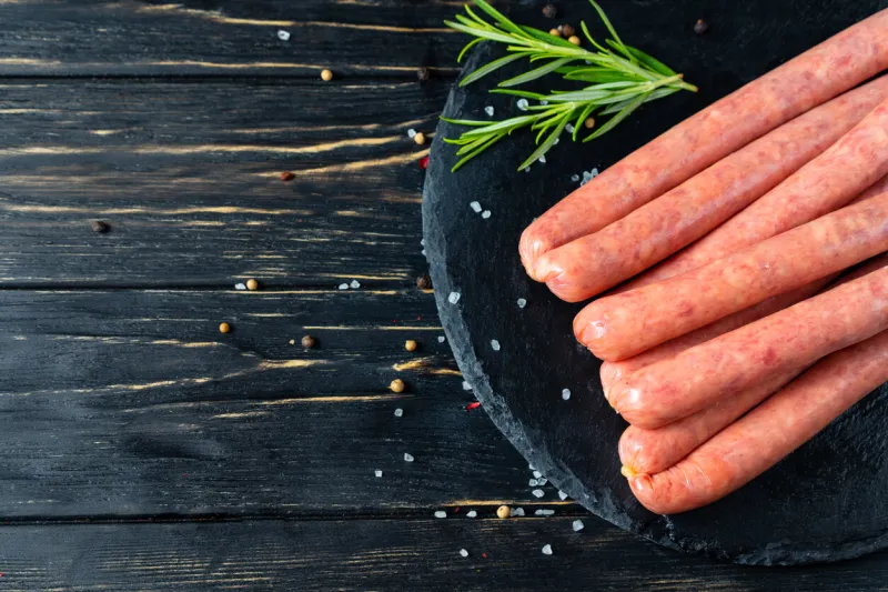 juicy sausages for barbecue lie on a stone chopping board with rosemary and spices, pepper and coarse salt on the black wooden table background
