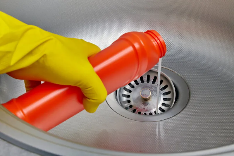 person's hand in a yellow rubber glove pours pipe cleaner down the drain of a metal kitchen sink prevention and cleaning of sewer blockages