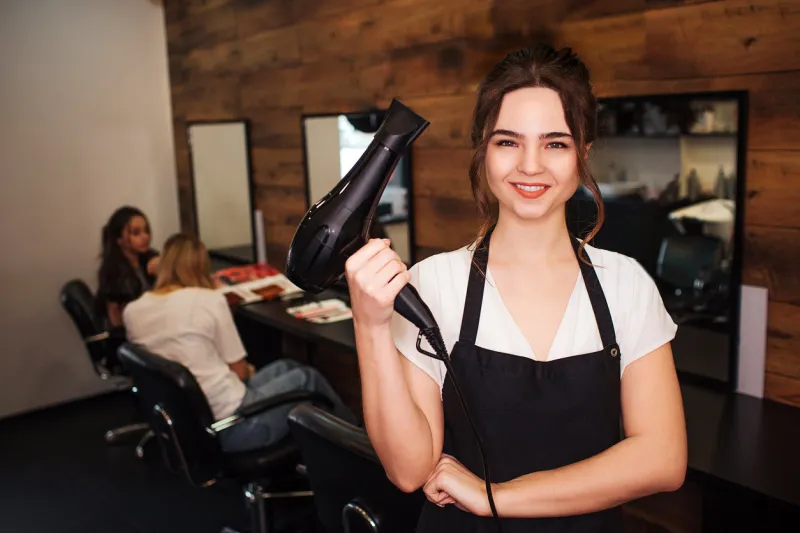 portret of smiling hairdresser in beauty salon beautiful woman in black apron looking at camera and holding professional black hair dryer beauty and people concept