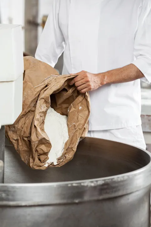 midsection of mature baker pouring flour in kneading machine at bakery