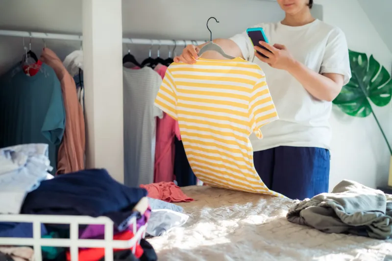 woman taking photo of t-shirt using her smartphone for selling or donating her clothes decluttering clothes, sorting, and cleaning up reuse, second-hand concept conscious consumer