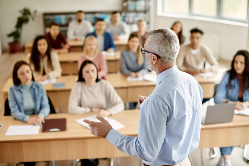 back view of mature professor giving lecture to large group of college students in the classroom