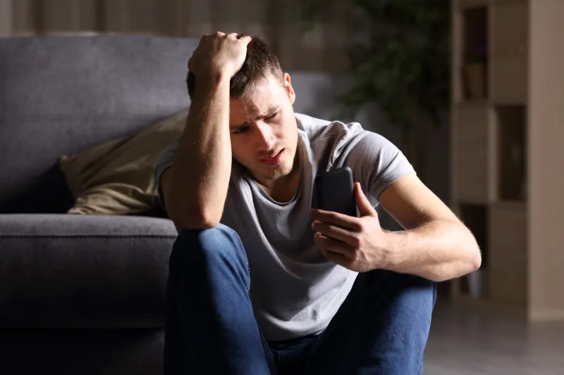 single sad man checking mobile phone sitting on the floor in the living room at home with a dark background