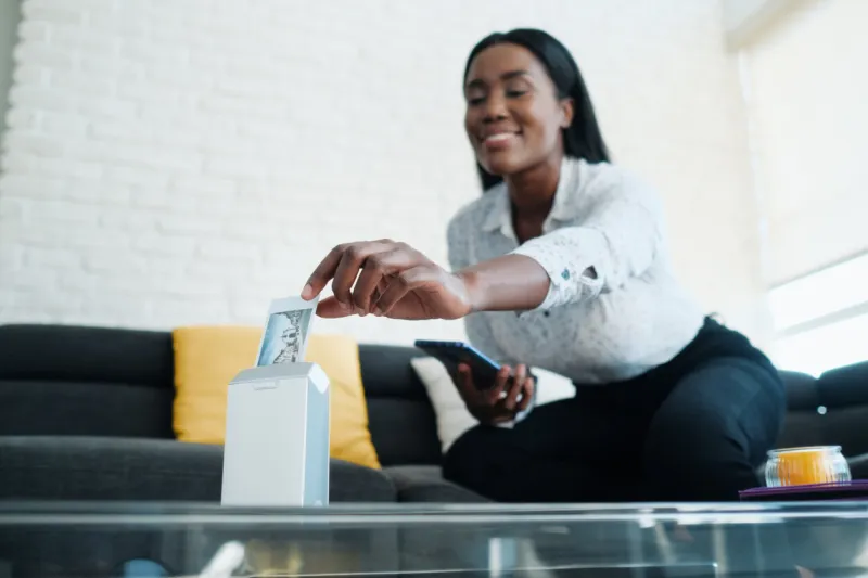 black woman printing pictures with portable wireless mini printer at home african american people sitting on sofa and archiving prints in photo album