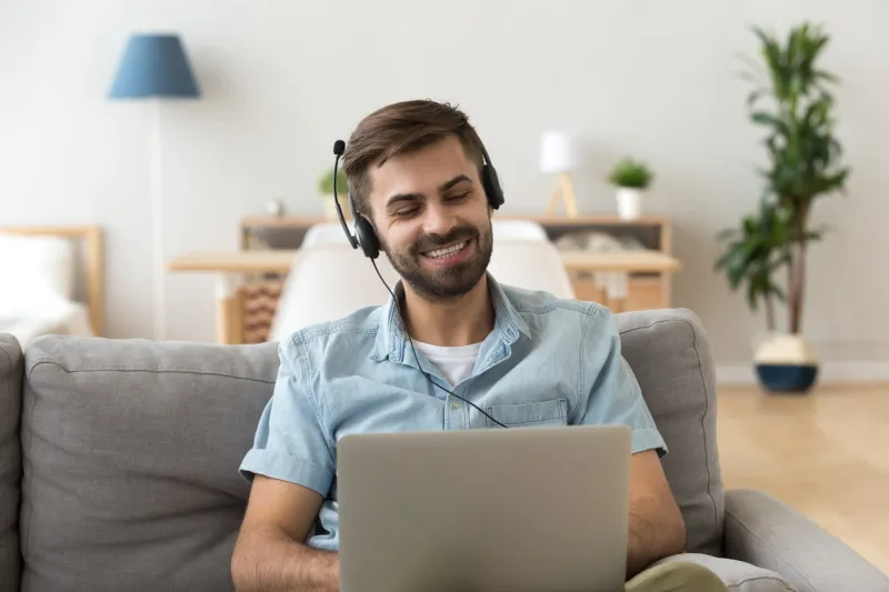 young happy man in headset sitting on couch at home looking at laptop studying online using computer, friendly guy watching webinar video, learning foreign language training, distance work education