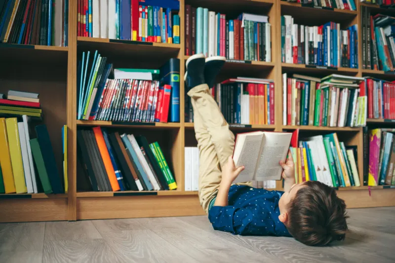 boy laying on the floor with the feet up, reading a book against multi colored bookshelf in library education, knowledge, bookstore, lecture pupil holds a book in his hands