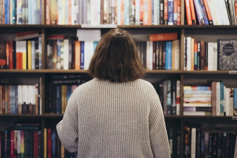 young brunette woman choosing a book in a bookstore, back view confused young woman seen from behind looking a the bookshelves thinking what book to buy at the bookstore