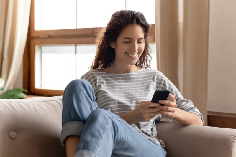 happy pleasant millennial woman relaxing on comfortable couch, holding smartphone in hands smiling young lady chatting in social networks, watching funny videos, using mobile applications at home