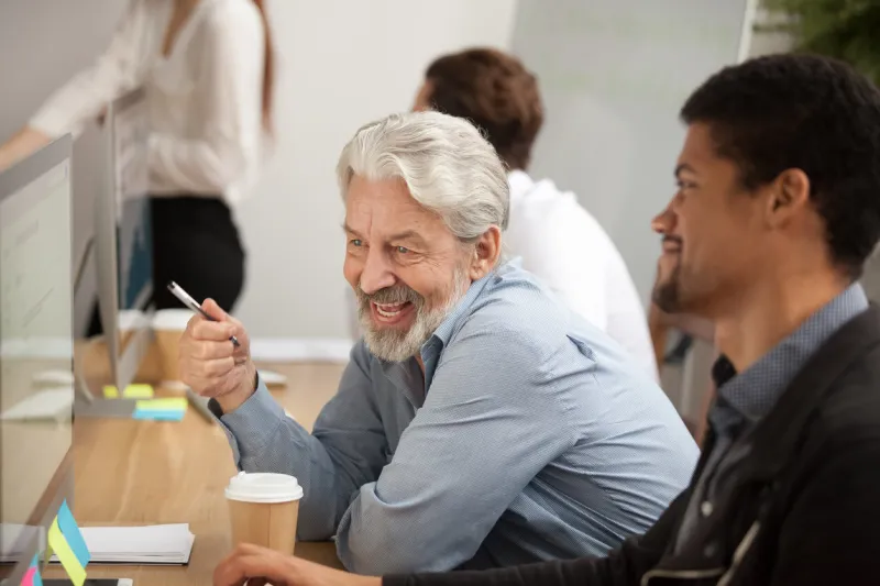 smiling senior employee discussing email with african colleague at workplace, happy older worker talking to black coworker joking about online computer work, aged and young managers laugh in office