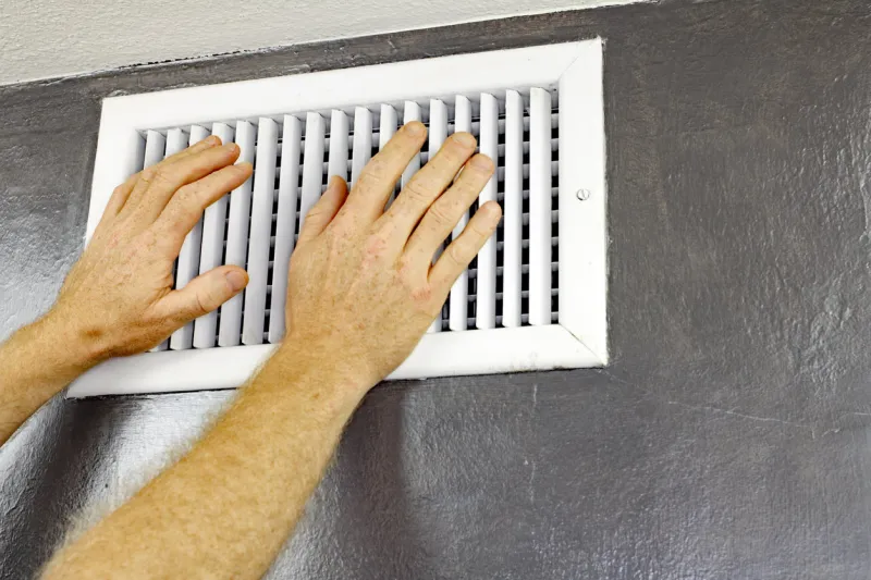 a pair of adult male hands feeling the flow of air coming out of an air vent on a wall near a ceiling man with hands in front of an air vent feeling for air flow