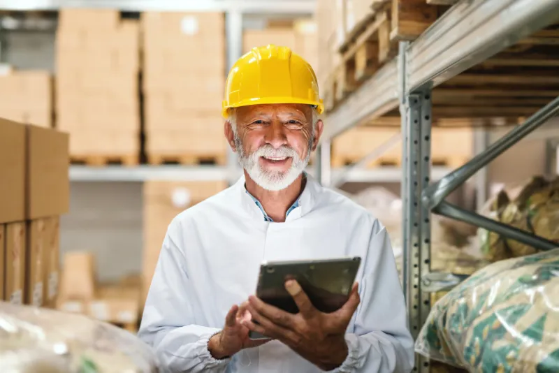 smiling senior worker in uniform and with yellow helmet on head holding tablet and looking at camera while standing in storage