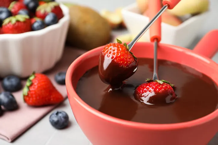 dipping strawberries into fondue pot with chocolate on table, closeup