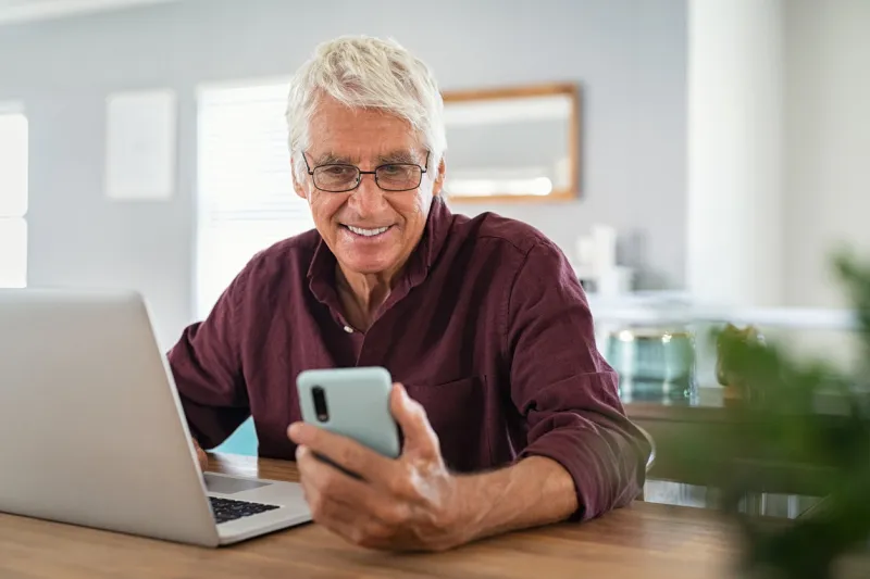 mature man using smartphone in front of laptop computer happy senior businessman working at home while messaging on mobile phone happy smiling old man using social media network technology and feeling excited