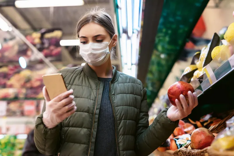 a young woman in a disposable face mask is checking a shopping list on a smartphone in a supermarket social distancing during the pandemic of the coronavirus covid-19