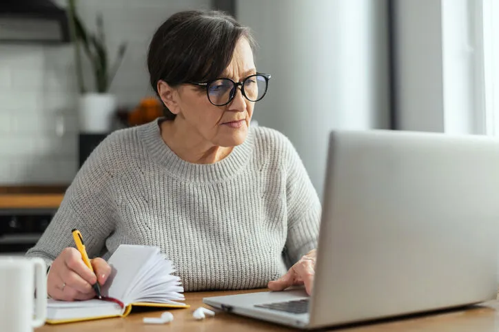 concentrated retirement woman using a laptop sitting at the desk in the kitchen for remote work, studying online, watching a webinar and takes a notes a senior business lady with computer indoor