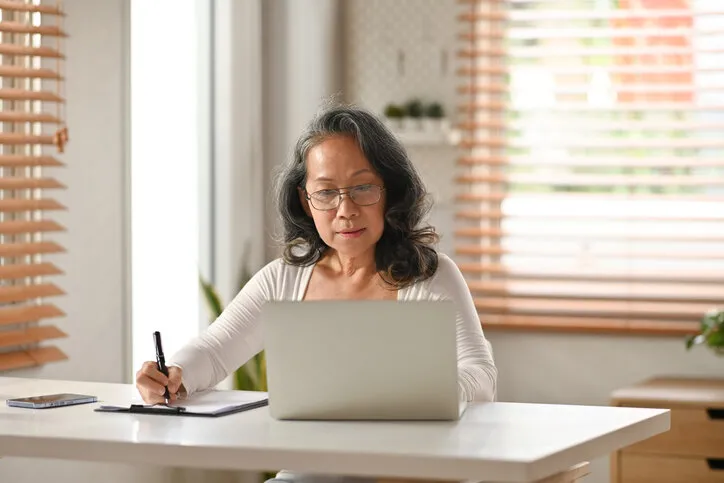 focused middle aged woman watching online webinar on laptop and making notes on notebook during remote working from home