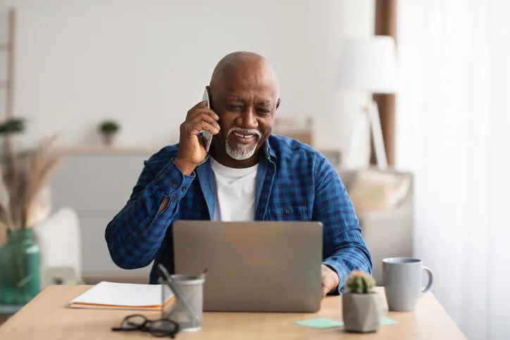 mature african american freelancer man talking on cellphone and using laptop computer sitting at desk in modern office senior businessman chatting via mobile phone business communication