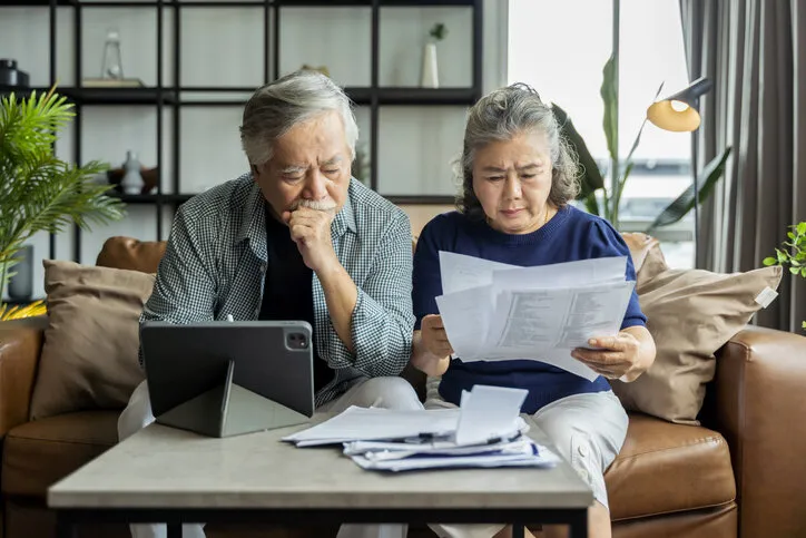 old retired asian senior couple checking and calculate financial billing together on sofa involved in financial paperwork, paying taxes online using e-banking laptop at living room home background