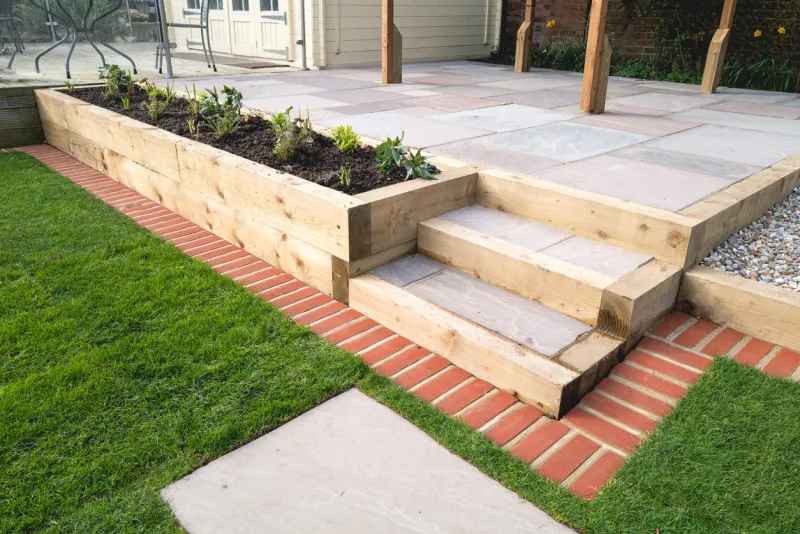 new steps in a garden or back yard leading to a raised patio, alongside a new raised flowerbed made using wooden sleepers a mowing strip of bricks is in front of newly laid turf