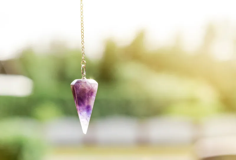 semi precious stone quartz pendulum on silver metal chain hanging outdoors in sunny day, green background