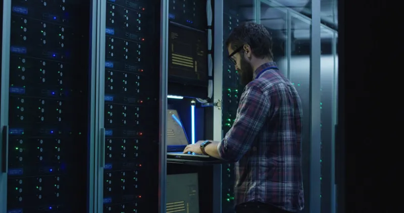 adult bearded system administrator in glasses walks over to the server rack and opens the rack door in the server room and makes the hardware settings using a laptop