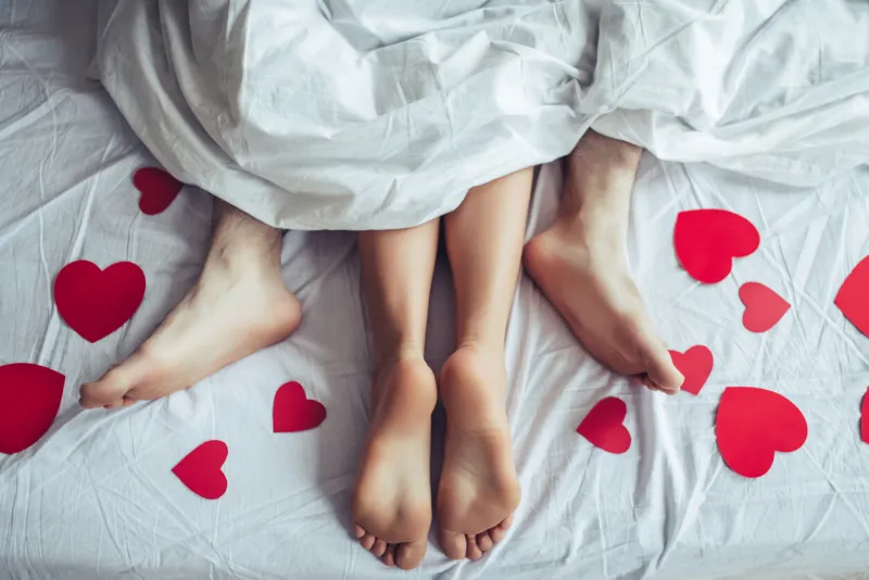 cropped image of young couple is lying on bed close up of male and female feet loving couple is lying on bed under blanket covered by small red paper hearts saint valentines day