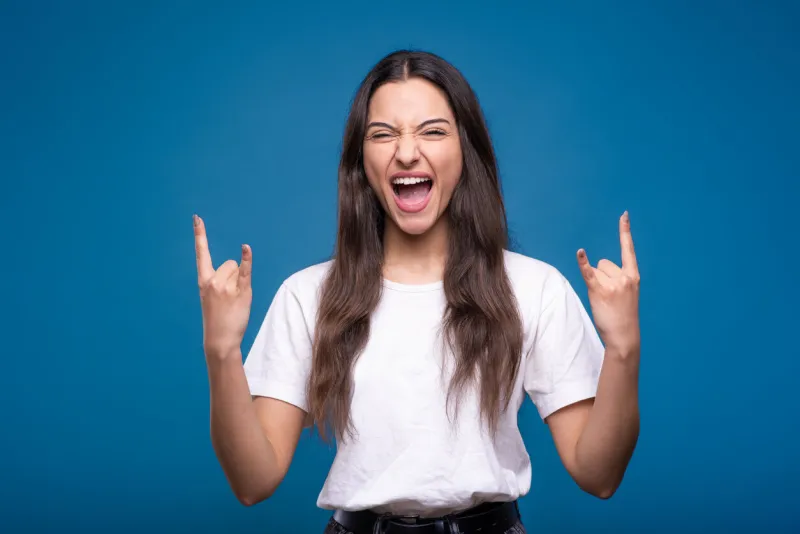 young and attractive caucasian or arab brunette girl in white t-shirt showing rocker and punk gesture with fingers and screaming isolated on blue studio background