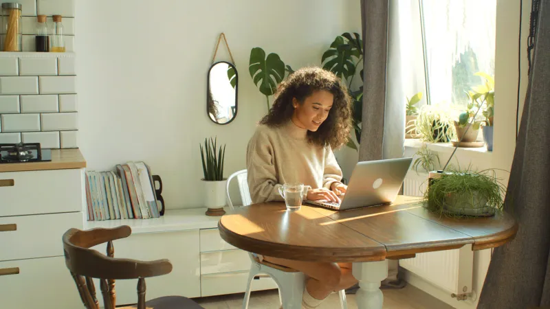 beautiful young brunette woman sitting in cozy kitchen by a table and working on her laptop computer