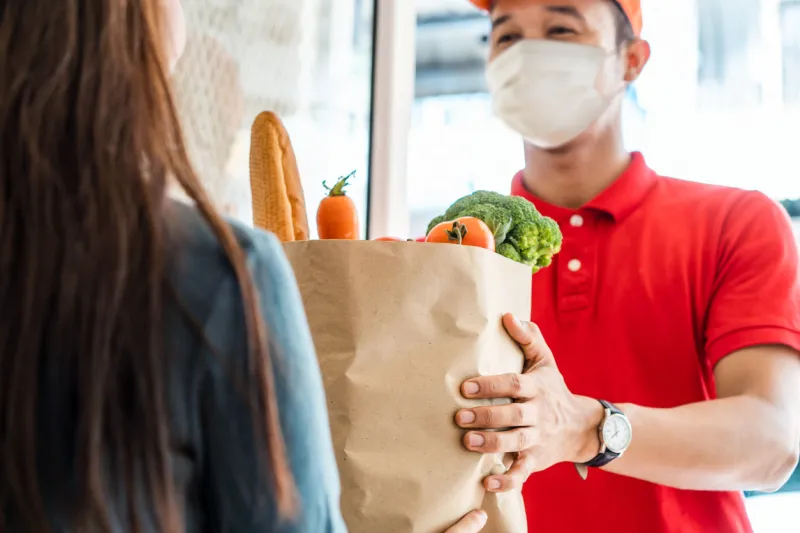 asian deliver man wearing face mask in red uniform handling bag of food, fruit, vegetable give to female costumer in front of the house postman and express grocery delivery service during covid19