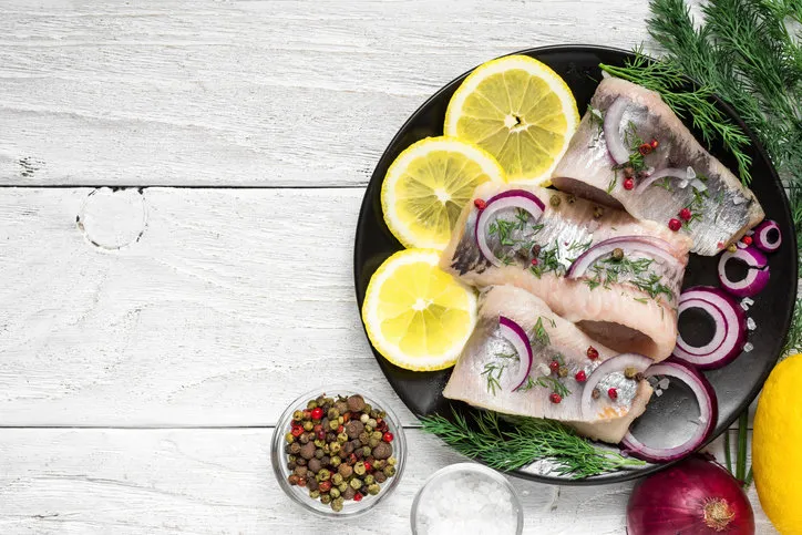 herring fillet with salt, pepper, herbs, onion and lemon on black plate with ingredients on white background top view healthy food
