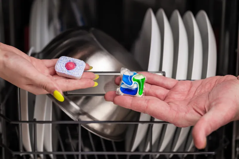 close-up of young woman and man hands holding two colored capsule for the dishwasher in the background, out of focus, is a dishwasher with clean dishes small depth of field