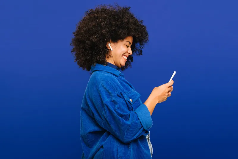 carefree young woman smiling happily while playing music using a smartphone and earbuds cheerful woman with curly hair enjoying her favourite playlist while standing against a blue background
