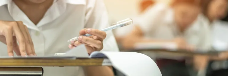 school student's taking educational exam, thinking hard, writing answer with left hand in classroom for education, university admission test and world literacy day concept