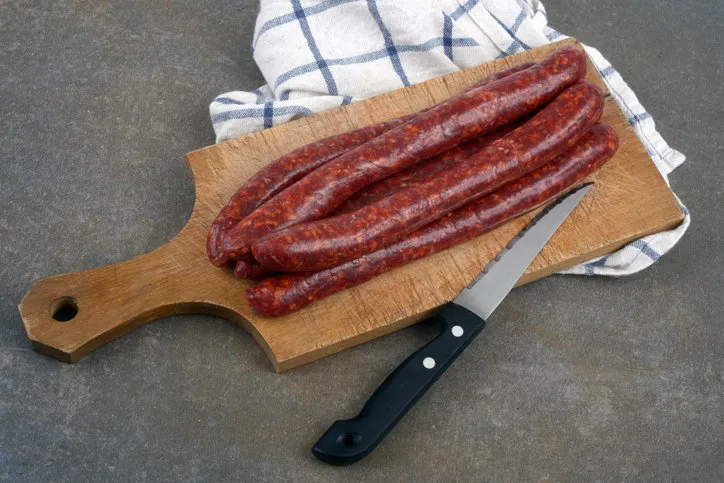 raw merguez on a wooden cutting board with a knife on a checkered cloth in close-up on a gray background