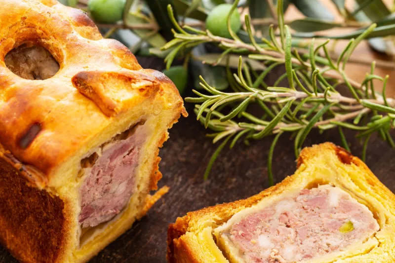 pate en croute or pâté en croûte or meat pie, with rosemary twig and green olives on branch with leaves over a dark wooden cutting board and a used oak wood background french traditional appetiser